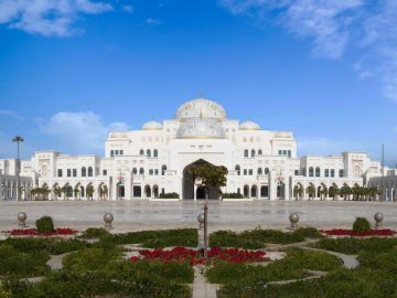 A wgite marble palace like structure under a blue sky in the background with green garden in the foreground