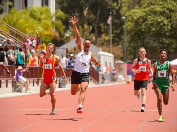 Athletes running on a red track with a winner in white vest rasing his one hand up in motion